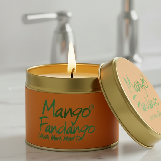 A lit candle in a round orange tin with a gold lid leaning against it on a light countertop. The label reads 'Mango Fandango' in green script, with a softly blurred bathroom sink and faucet in the background.