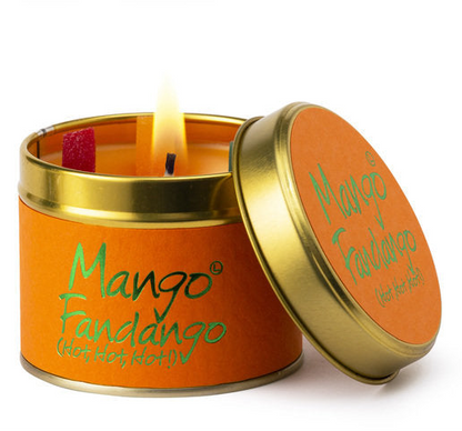 A lit orange candle in a round gold metal tin with the lid leaning against it. The label reads 'Mango Fandango (Hot Hot Hot!)' in green script, and a small red wax piece is visible inside the candle.