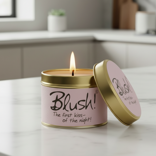 A pink scented candle in a gold metal tin sits on a light marble countertop with the lid leaning against the front. The candle is lit, and the label reads 'Blush! The first kiss- of the night!'.