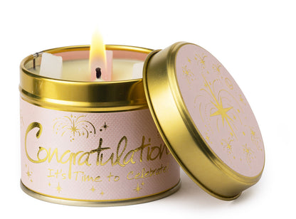A lit candle in a round gold metal tin with a pink label that reads 'Congratulations It's Time to Celebrate' in gold script and stars, shown on a white background with the lid leaning against the tin.