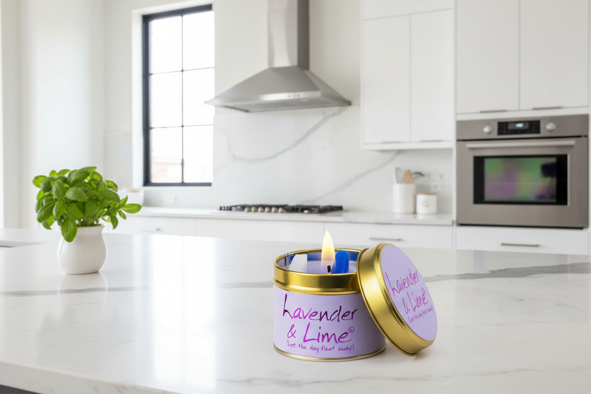 A lavender and lime scented candle in a round tin with a gold lid, set on a white marble kitchen countertop with a lit flame. The background shows a modern white kitchen with a range hood and oven.
