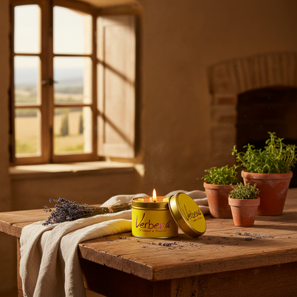 A yellow cylindrical candle tin with the brand name 'Verbena' on the lid, lit and placed on a rustic wooden table in a warm, sunlit room. Lavender stems, potted herbs, and a draped beige cloth surround it, with a blurred window and fireplace in the background.