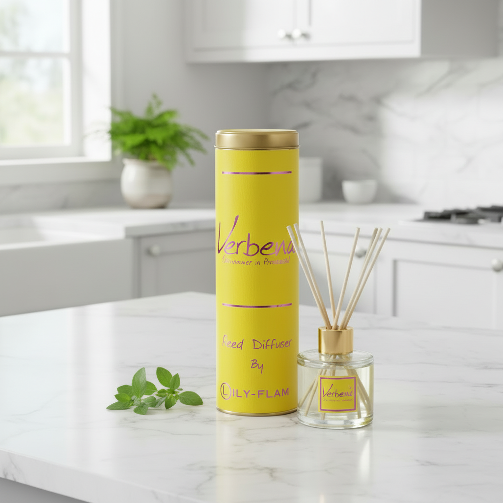 A yellow reed diffuser set on a white marble kitchen counter, featuring a tall cylindrical yellow container with a gold lid and a matching glass reed diffuser bottle with wooden reeds. The background shows a softly blurred modern kitchen with white cabinets and a potted plant.