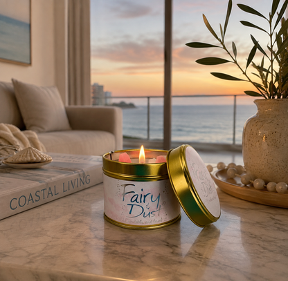 A lit tin candle labeled 'Fairy Daze' sits on a marble table in a coastal living room setting with a sunset over the ocean, a sofa, and a potted olive tree in the background.