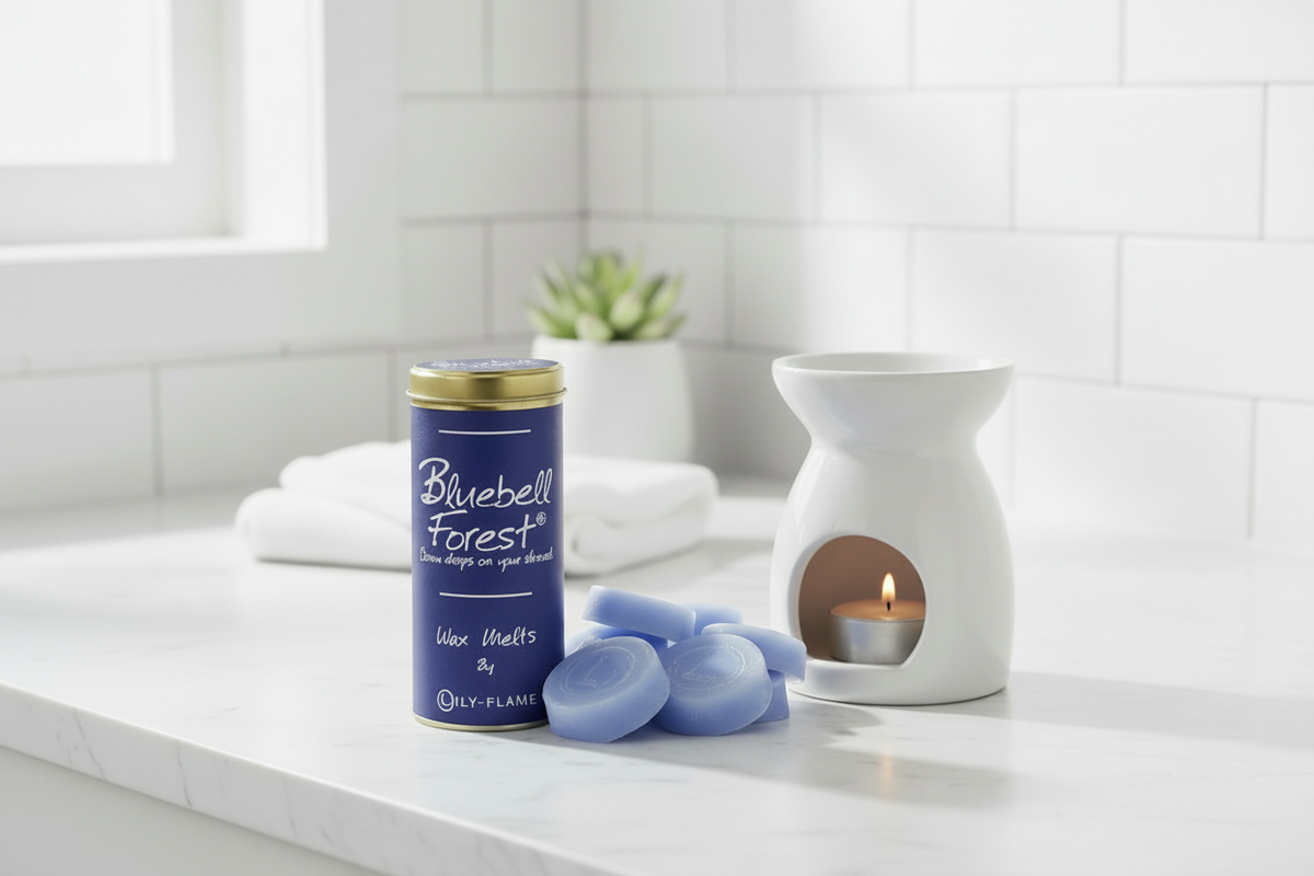 A bathroom or spa-like setting with a blue cylindrical candle tin labeled Bluebell Forest, several pale blue wax melts, a white ceramic candle warmer with a lit candle, folded white towels, and a small potted succulent on a light marble counter.