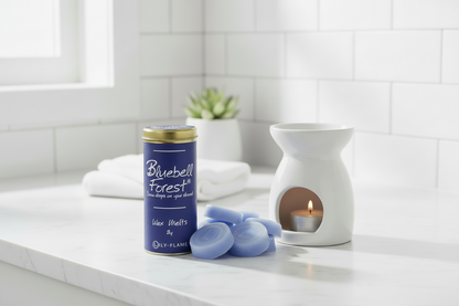 A bathroom or spa-like setting with a blue cylindrical candle tin labeled Bluebell Forest, several pale blue wax melts, a white ceramic candle warmer with a lit candle, folded white towels, and a small potted succulent on a light marble counter.