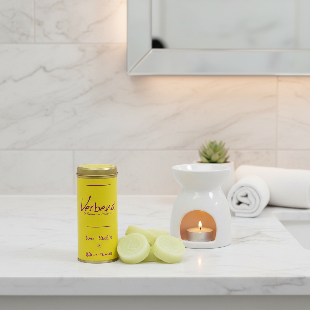 A yellow tin of Verbena room fragrance sits on a white marble bathroom counter next to a white ceramic candle warmer with a lit tealight candle, with rolled towels and a mirror in the background.
