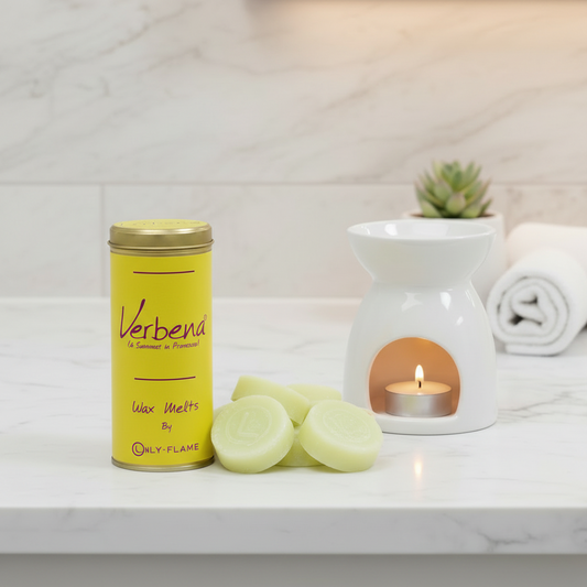 A yellow tin of Only Flame Verbend Wax Melt in a scented floral scent sits on a white marble countertop next to a white ceramic candle warmer with a tea light candle lit inside. Fresh pale green wax melts are scattered in front, with a small potted succulent and a rolled towel blurred in the background.
