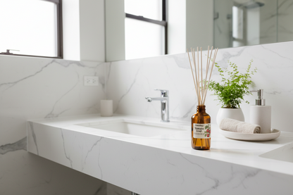 An amber glass reed diffuser bottle with wooden reeds sits on a white marble-look bathroom counter next to a chrome faucet, a small potted green plant, and a white soap dispenser.