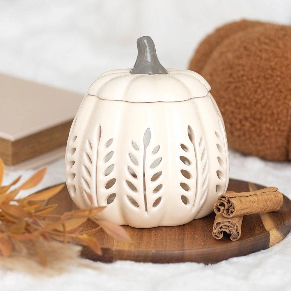 White ceramic pumpkin-shaped lantern with cut-out patterns on a wooden surface with cinnamon sticks and leaves.