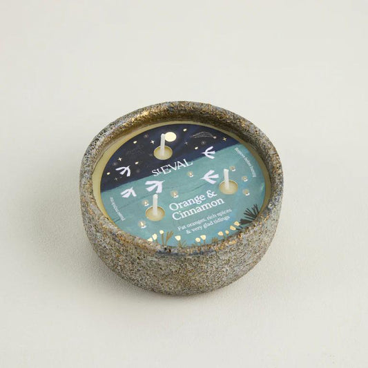 Candle in a textured holder with a label indicating 'Orange & Cinnamon' fragrance on a light background.