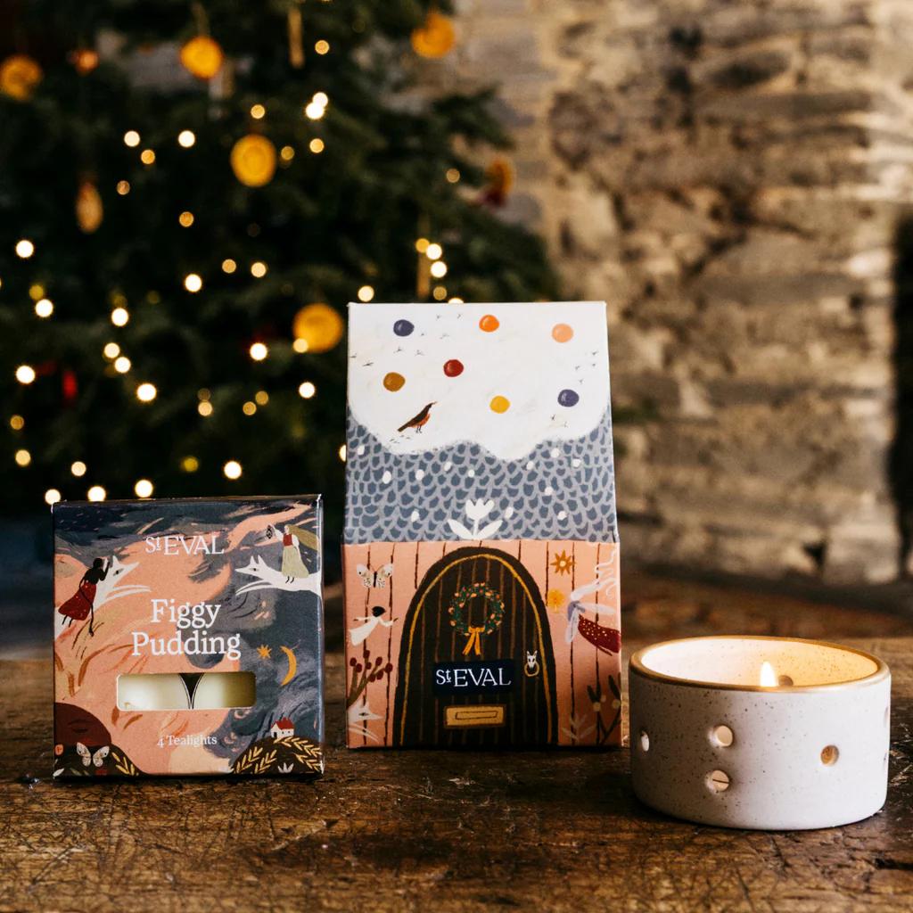 ST Eval Figgy Pudding candles with decorative packaging on a wooden surface in front of a Christmas tree.