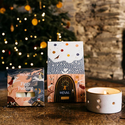 ST Eval Figgy Pudding candles with decorative packaging on a wooden surface in front of a Christmas tree.