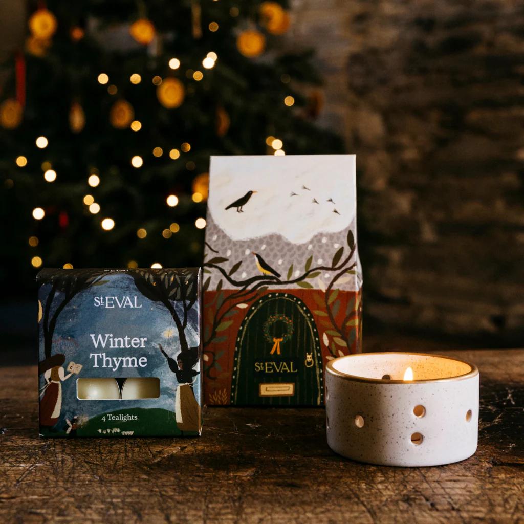 Two candle boxes and a lit candle on a wooden surface with a Christmas tree in the background.