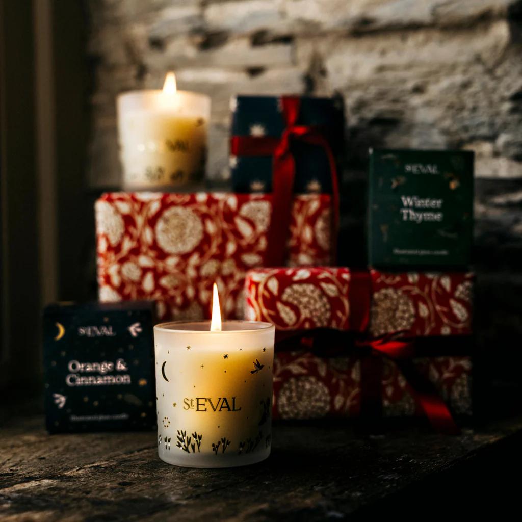 Candle with 'St EVAL' branding in front of wrapped gifts and another candle.