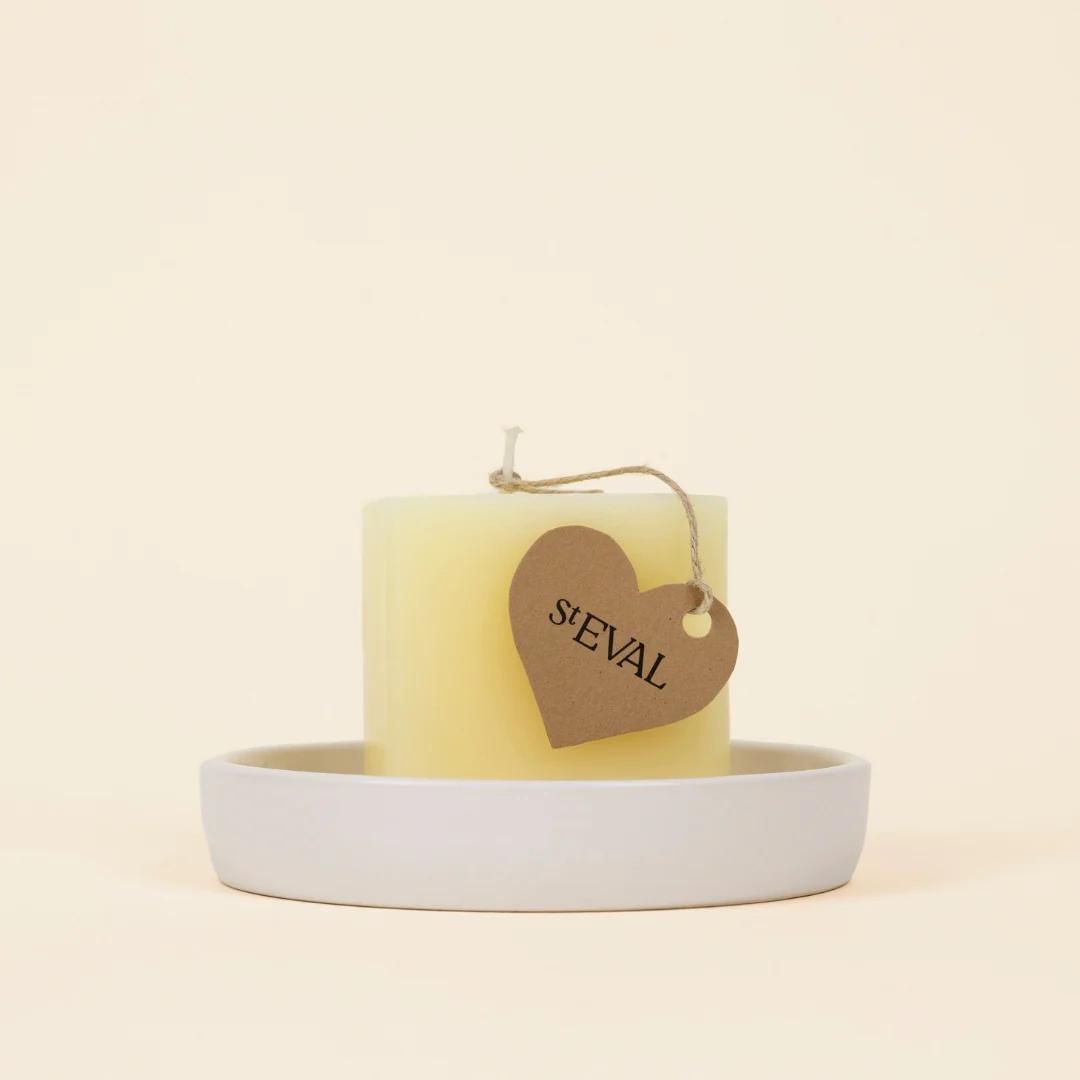 Yellow candle with a heart-shaped tag labeled 'St EVAL' on a white dish against a beige background