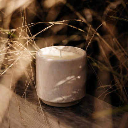 Ceramic candle holder with a candle inside, surrounded by dry grass on a wooden surface.