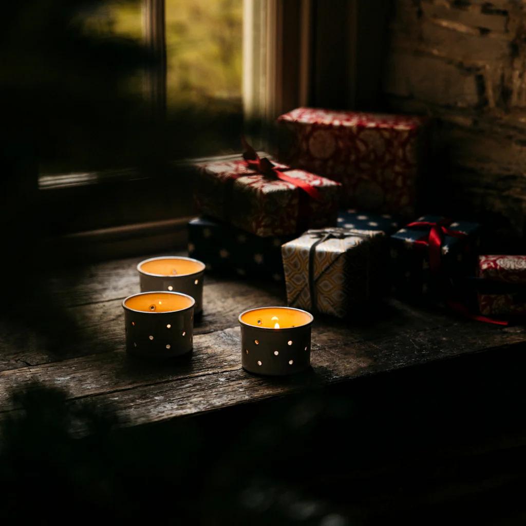 Three lit candles in black holders on a wooden surface with wrapped gifts in the background.