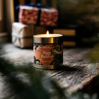 Candle labeled 'Figgy Pudding' by ST EVAL on a wooden surface with gift boxes in the background.