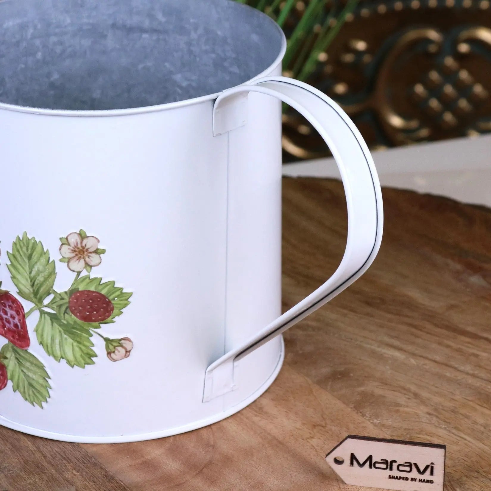 White metal watering can with floral design on a wooden surface, Maravi brand visible.