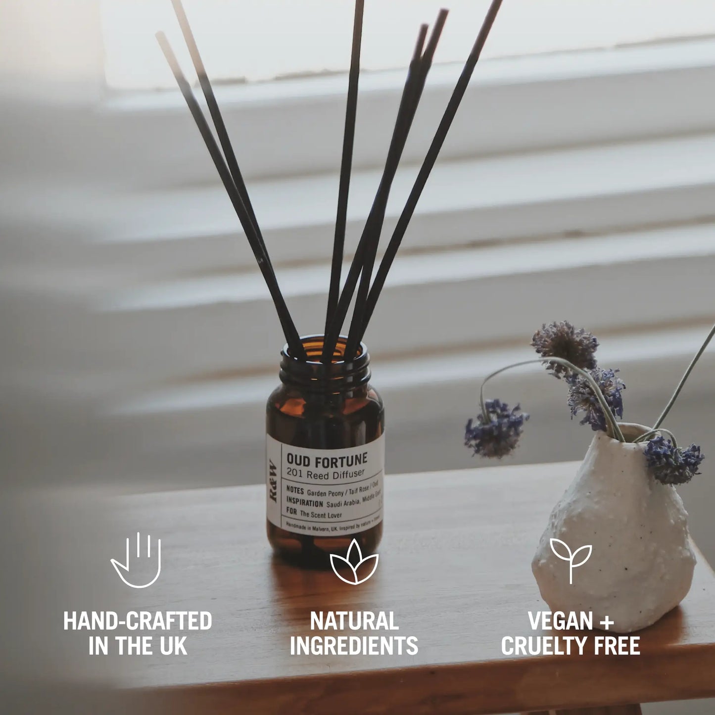 Reed diffuser with decorative vase on a wooden surface, featuring icons and text about product attributes.