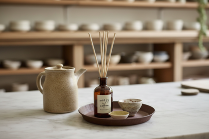 A reed diffuser bottle with wooden reeds on a round tray, styled on a marble countertop with a ceramic teapot and bowls in a warm, softly blurred kitchen setting.