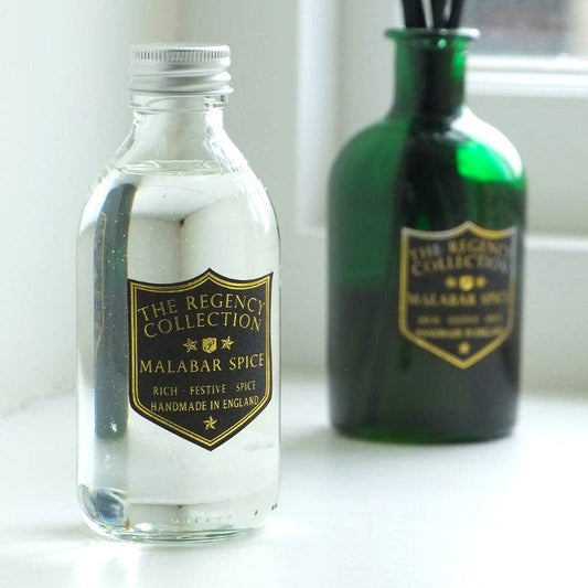 A clear glass bottle of Malabar Spice from The Regency Collection with a gold label and silver cap, standing in front of a green glass reed diffuser on a white surface.