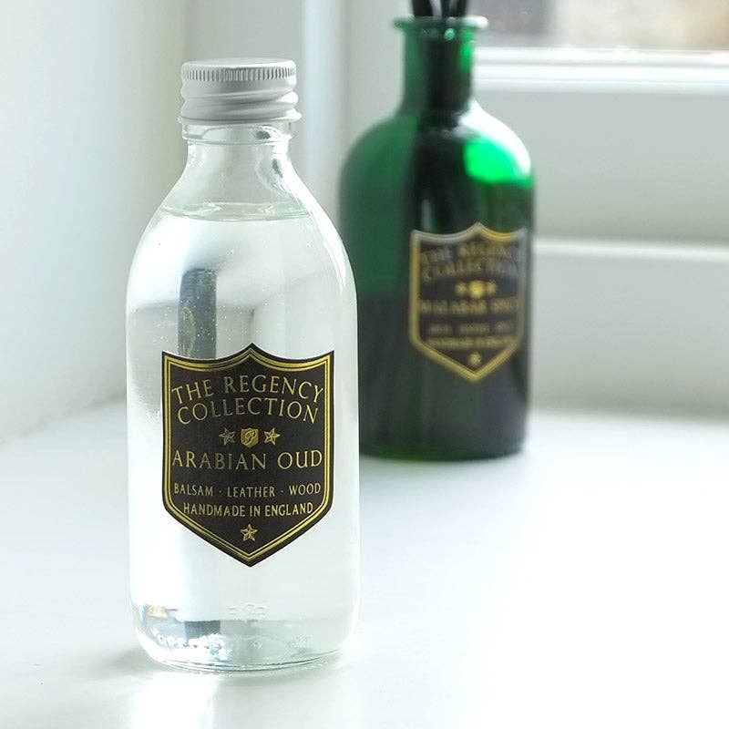 A clear glass bottle of Arabian Oud balsam from The Regency Collection in the foreground, with a gold shield label, set on a white surface next to a blurred green bottle in the background.