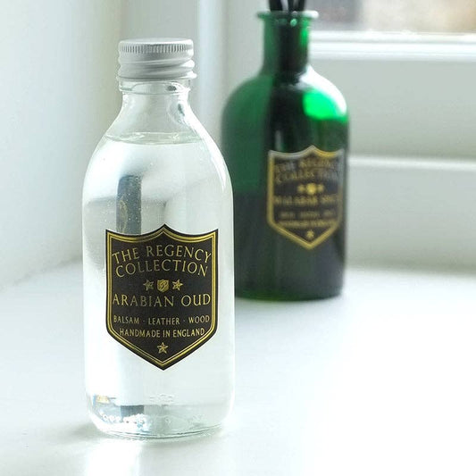 A clear glass bottle of Arabian Oud balsam from The Regency Collection in the foreground, with a gold shield label, set on a white surface next to a blurred green bottle in the background.