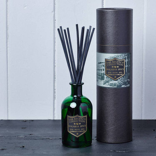 A reed diffuser bottle made of dark green glass with black reeds, standing next to its tall dark gray cylindrical packaging tube. Both pieces feature a gold and white label for 'The Regency Collection' and 'Palafrina Spice'.