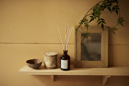A reed diffuser bottle with wooden reeds stands on a light wood shelf against a warm yellow wall, styled with a ceramic bowl, a textured tin, and a framed mirror, with a green leafy vine hanging from above.